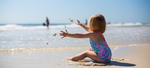 A little girl sitting on the beach in shallow water playing with sand.